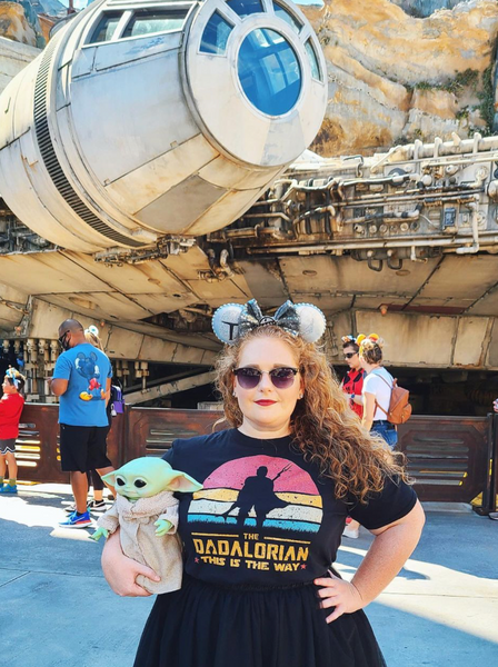 The Dadalorian Shirt This Is The Way the mandalorian mando daddy zaddy pedro pascal Star Wars dad fathers day gift outfit disney dad DILF outfit cosplay man men cotton unisex women baby yoda grogu tshirt graphic tee comic con Woman Wearing shirt at Disney World Hollywood Studios Galaxy's Edge holding grogu puppet doll with mandalorian mickey ears and sunglasses in front of the Millennium Falcon smugglers run ride