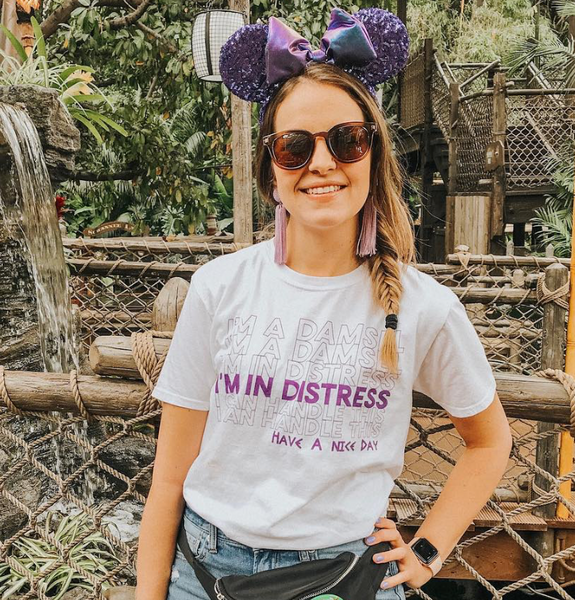 Woman wearing Meg Hercules I'm A Damsel I'm In Distress I Can Handle This Have A Nice Day T-Shirt, unisex graphic tee, disneyland disney world vacation matching shirts, hipster disney princess, megara I can go the distance. Size inclusive XS-4XL in Red and Purple. Thank you Have a nice day. Purple earrings Purple Potion Mickey Ears, photo in front of the Jungle Cruise Ride by the Dole Whip Stand.