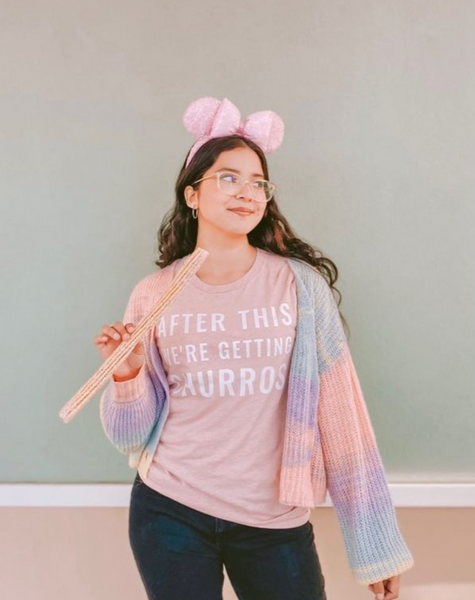 Woman wearing After This We're Getting Churros Shirt in Pink with mickey mouse minnie ears rose gold holding Disneyland churro food snack and lover pink blue purple tie dye cardigan with white glasses and gold hoop earrings jewelry