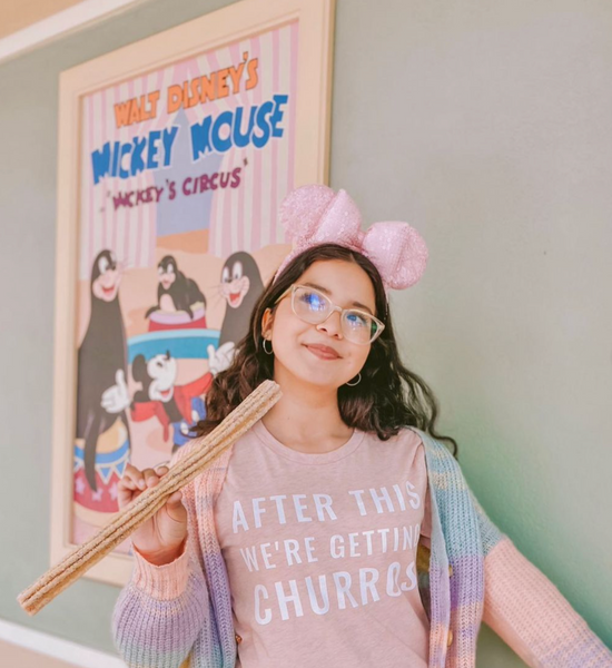 Woman wearing After This We're Getting Churros Shirt in Pink with mickey mouse minnie ears rose gold holding Disneyland churro food snack and lover pink blue purple tie dye cardigan with white glasses and gold hoop earrings jewelry. Walt Disney's Mickey Mouse Circus Poster in background.
