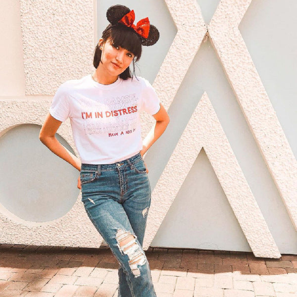 Woman wearing Meg Hercules I'm A Damsel I'm In Distress I Can Handle This Have A Nice Day T-Shirt, unisex graphic tee, disneyland disney world vacation matching shirts, hipster disney princess, megara I can go the distance. Size inclusive XS-4XL in Red and Purple. Thank you Have a nice day. Red Minnie Ears photo in front of the Small World Wall at Disneyland Anaheim California.