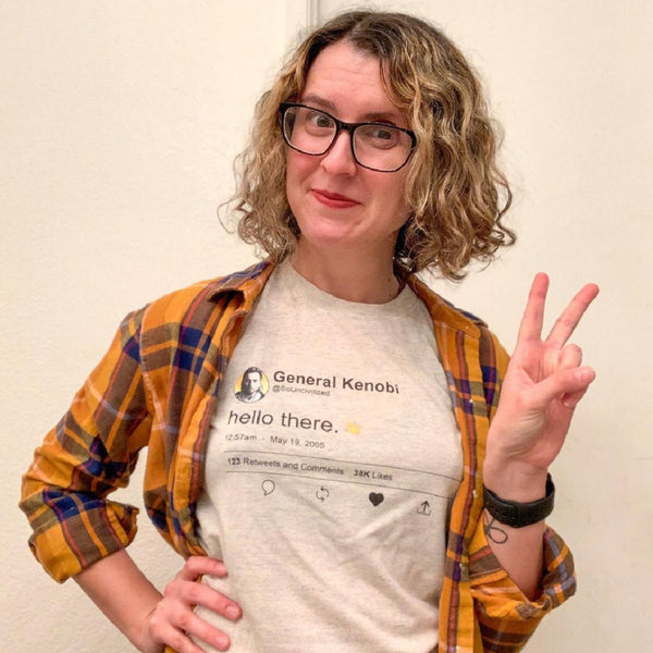 Woman with curly hair wearing General Kenobi Hello There tweet shirt with wave hey hello hand emoji. Prequels Easter eggs, may 19 2005, attack of the clones, general grievous, so uncivilized Obi Wan movie quotes. Available in Sand Beige and Black, size inclusive XS-4XL. Worldwide shipping. Making a peace sign with hand and wearing black glasses and watch.