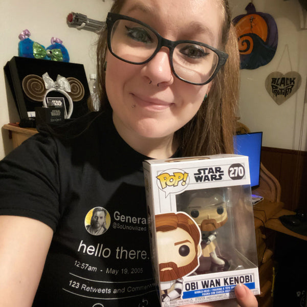 Woman at home wearing General Kenobi Hello There tweet shirt with wave hey hello hand emoji. Prequels Easter eggs, may 19 2005, attack of the clones, general grievous, so uncivilized Obi Wan movie quotes. Available in Sand Beige and Black, size inclusive XS-4XL. Worldwide shipping. Holding funko pop obi wan kenobi toy merch wearing black glasses