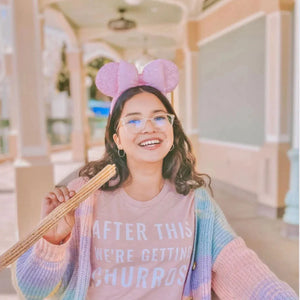 Picture of woman wearing After This We're Getting Churros Shirt in Rose Gold by Shop Friday Apparel at Disneyland Park eating a churro wearing Mickey Minnie Ears in Pink with Rainbow Cardigan Sweater