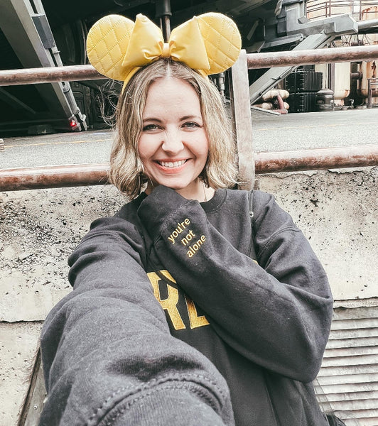 Woman wearing embroidered sweatshirt that says REYLO on the front and you're not alone on the left sleeve. At Galaxy's Edge Batuu by Kylo Ren's tiefighter. For Star Wars fans of Kylo Ren, Ben Solo and Rey. Shown in black with yellow embroidery.