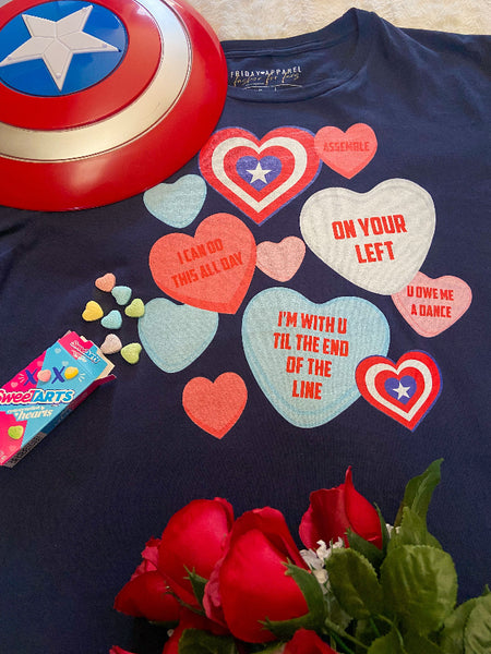 Flatlay of Captain America Candy Hearts Conversation Quote T-Shirt. Navy gildan softstyle graphic tee with red, blue, pink, white hearts and quotes from Avengers Marvel movies. Assemble, on your left, i can do this all day, i'm with you til the end of the line, you owe me a dance. Valentine's Day. Avenger's Campus. Sweetarts candy and Captain America shield with red roses