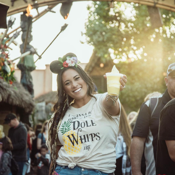 Woman wearing I Wanna Be Where The Dole Whips Are Shirt in cream with black design and yellow pineapple holding a Disneyland Dole Whip mickey food snack. Wearing aloha floral mickey mouse ears.