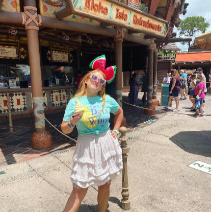 Woman wearing I Wanna Be Where The Dole Whips Are Shirt in mint green with purple design and yellow pineapple holding a Walt Disney World Dole Whip mickey food snack. Wearing watermelon giant mickey mouse ears and white skirt with white shoes and yellow pineapple mini backpack loungefly.