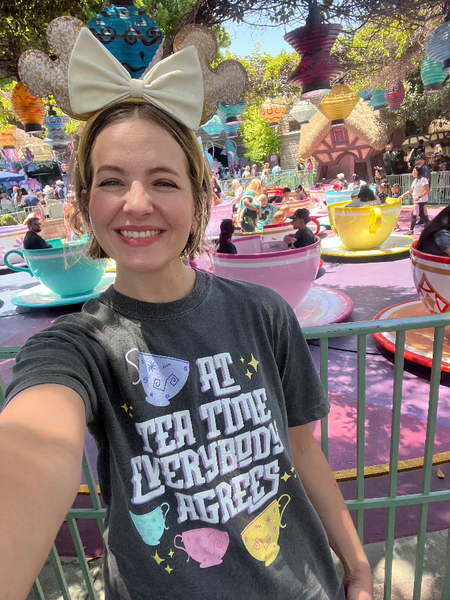 Woman wearing At Tea Time Everybody Agrees Shirt Comfort Colors Grey with Purple, Teal Blue, Pink and Yellow teacups ride design. Disney Swifties Outfit Top Eras Tour Midnights Music Lyrics Theme Park. At Disneyland Teacups Ride wearing Beignet All Day Mickey Ears food snacks