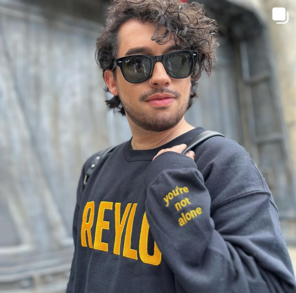 Man wearing embroidered sweatshirt that says REYLO on the front and you're not alone on the left sleeve with black sunglasses. For Star Wars fans of Kylo Ren, Ben Solo and Rey. Shown in black with yellow embroidery. At Galaxy's Edge Batuu at disneyland california.