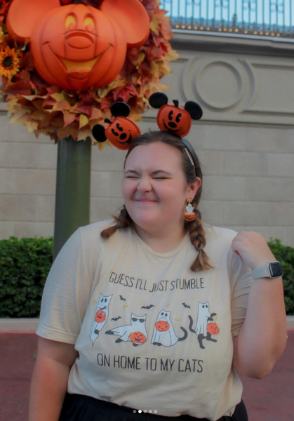 Woman at Disney Halloween wearing beige ‘Guess I’ll Just Stumble on Home to My Cats’ t-shirt with ghost cats and jack-o-lanterns, styled with Mickey pumpkin ears and ghost earrings.