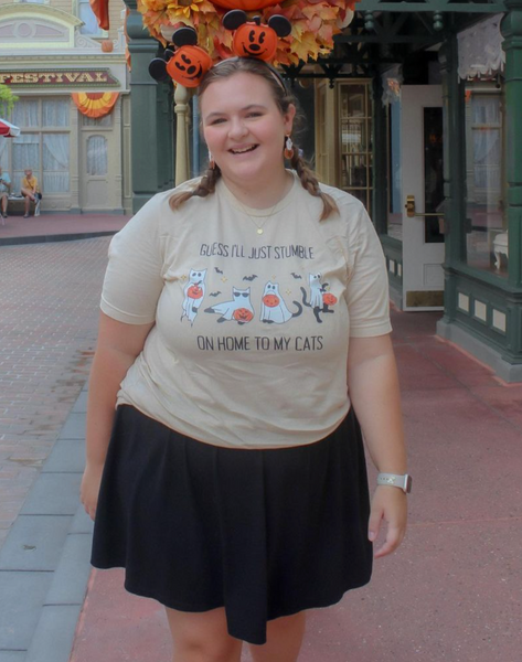 Woman at Disney Halloween wearing beige ‘Guess I’ll Just Stumble on Home to My Cats’ t-shirt with ghost cats and jack-o-lanterns, styled with Mickey pumpkin ears and ghost earrings.
