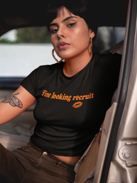 Woman wearing a black unisex graphic t-shirt with distressed orange text reading “Fine Looking Recruit” and a small orange kiss graphic.