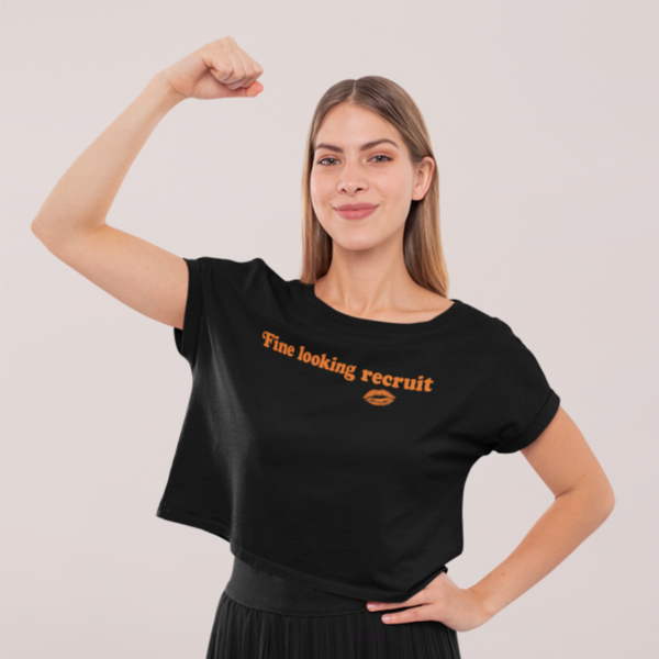 Woman wearing a black unisex graphic t-shirt with distressed orange text reading “Fine Looking Recruit” and a small orange kiss graphic.