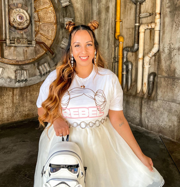 Woman in a white dress with 'REBEL' print, holding a stormtrooper helmet in a themed setting.