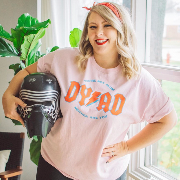 Woman wearing a pink Dyad in the Force t-shirt with a colorful graphic and text, holding a Kylo Ren helmet indoors.
