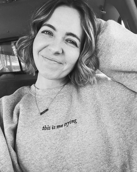 Woman wearing embroidered sweatshirt in grey that says this is me trying from folklore taylor swift music album music lyrics quote. Black embroidery on gray fabric. Long sleeve pullover sweater. Woman with short curly hair and necklace. Inspirational outfit smiling photo.