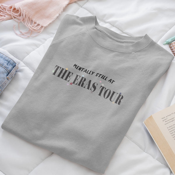 Flatlay of Mentally Still At The Eras Tour Sweatshirt colorful star for each era taylor swift swiftie sweater grey unisex cotton polyester top books on bed with jeans and pink scarf with planner notebook journal and mint colored pen