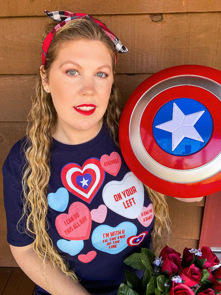 Woman wearing Captain America Candy Hearts Conversation Quote T-Shirt. Navy gildan softstyle graphic tee with red, blue, pink, white hearts and quotes from Avengers Marvel movies. Assemble, on your left, i can do this all day, i'm with you til the end of the line, you owe me a dance. Valentine's Day. Avenger's Campus. Plaid black red headband with red lipstick and Captain America shield.