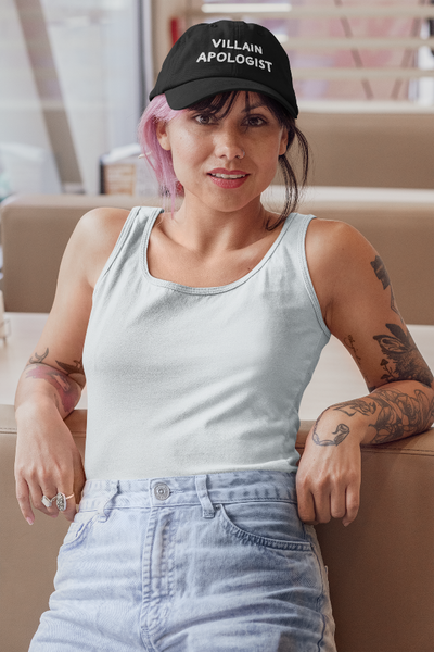 Woman wearing black Villain Apologist embroidered baseball cap. Black grunge bleached wash fabric with white embroidered thread design. Light blue tank top with arm tattoos and finger rings and pink short hair.