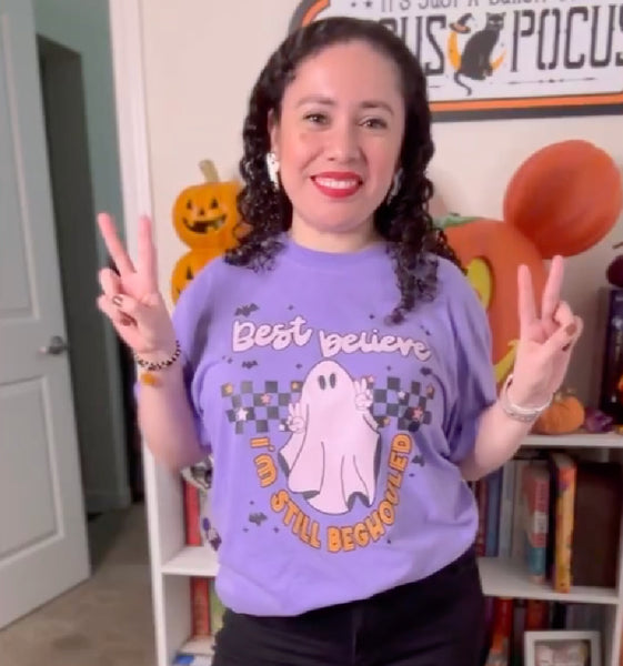 Purple graphic t-shirt with pink ghost making peace signs, black and white checkerboard background, bats, stars, and text ‘Best believe I’m still beghouled’ – cute Halloween Taylor Swift inspired tee. bejeweled midnights graphic tee taylor travis engaged purple woman pumpkin smile costume