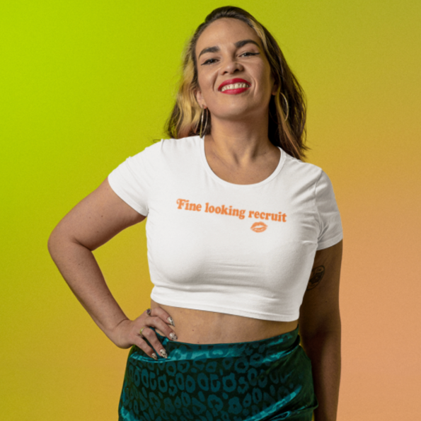 Woman wearing a white unisex graphic t-shirt with distressed orange text reading “Fine Looking Recruit” and a small orange kiss graphic. cropped tee