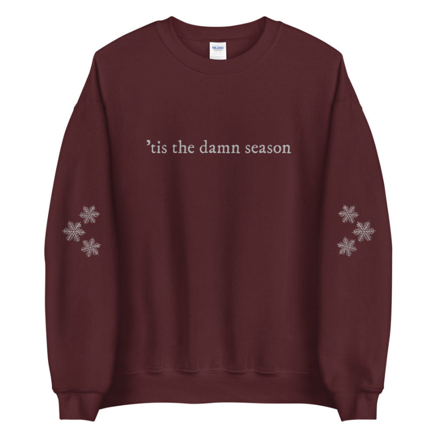 Burgundy unisex sweatshirt with embroidered text ’tis the damn season and snowflake sleeve embroidery; Swiftie-inspired winter crewneck.