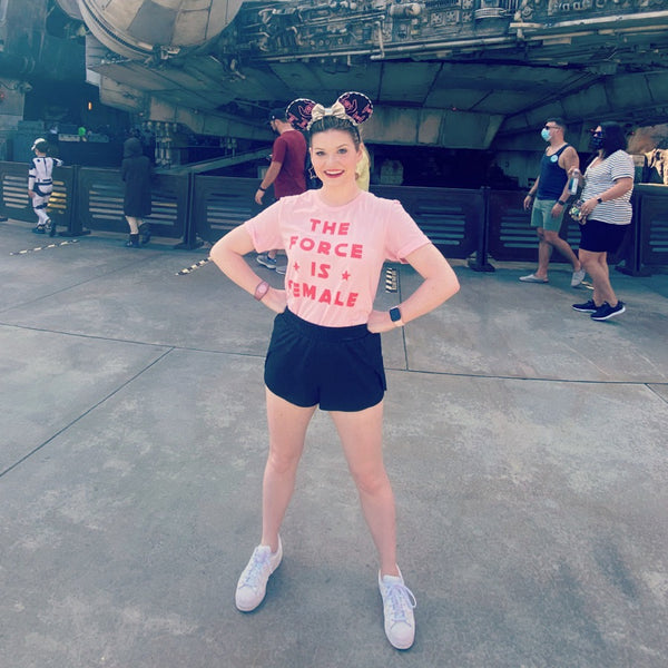 The Force Is Female Star Wars inspired girl power shirt in White with Black print and stars at Galaxy's Edge Batuu bound outfit in front of Smuggler's Run Millennium Falcon. Style with a skirt and high top combat boots and Mickey ears. Disneyland California and Hollywood Studios Orlando Florida. Size inclusive XS-4XL. Worldwide shipping.